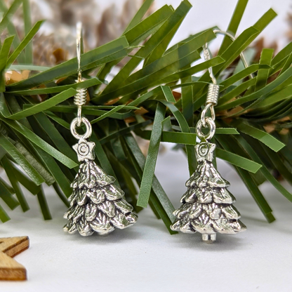 A pair of Silver Sunbird 3D Christmas Tree Earrings are displayed in front of artificial green pine branches, with a small wooden star in the bottom left corner.