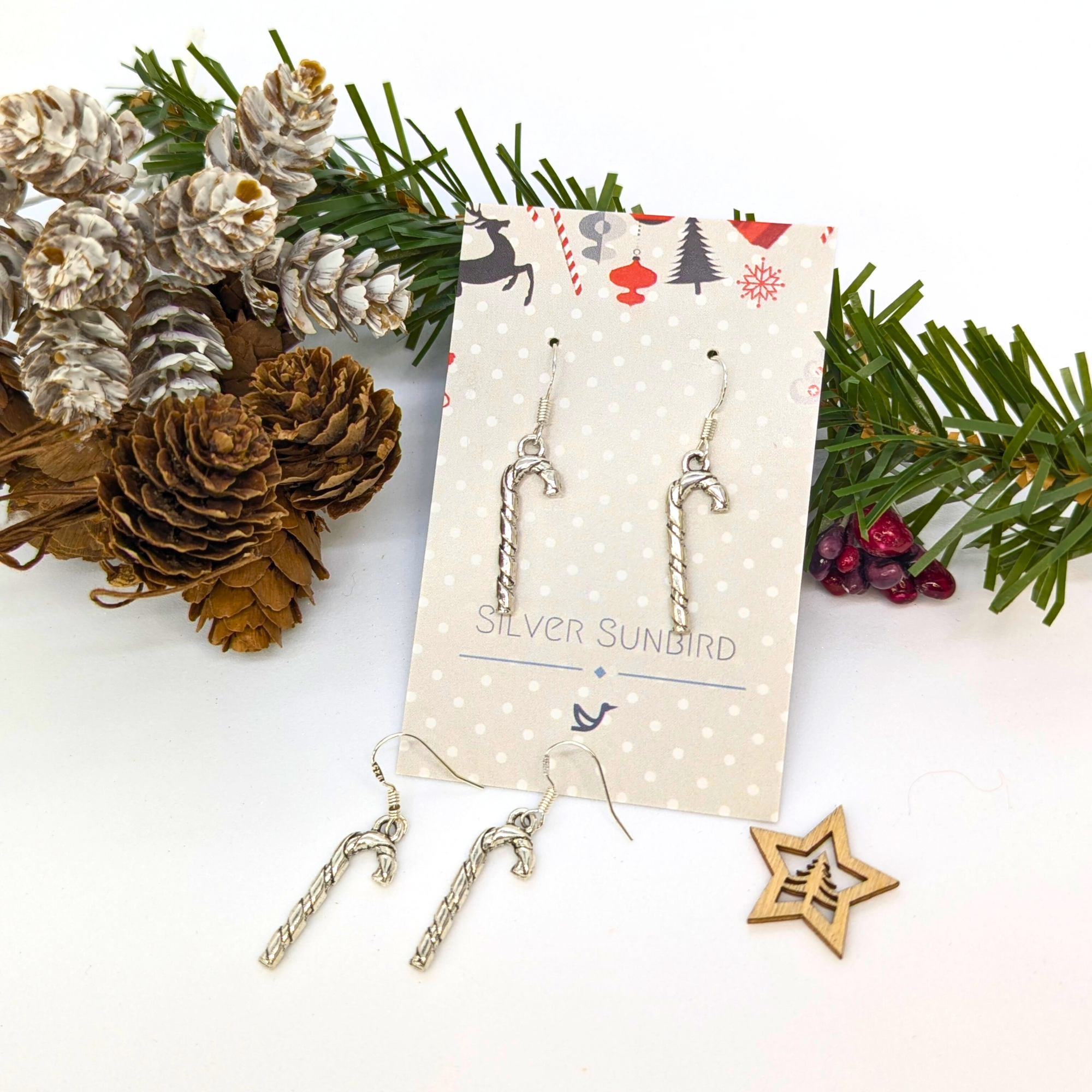 Classic Candy Cane Earrings by Silver Sunbird in sterling silver, displayed on a festive holiday card beside pinecones, greenery, red berries, and a wooden star—the perfect Christmas accessory.