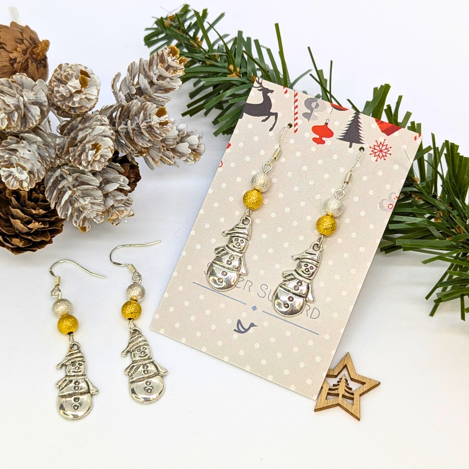 Very Merry Snowmen Earrings by Silver Sunbird—festive, handmade sterling silver earrings with gold bead accents, presented on a decorative card and artfully arranged with pine branches, pine cones, and a wooden star.