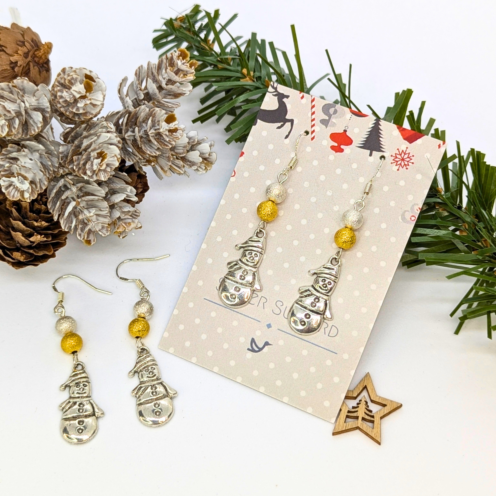 Very Merry Snowmen Earrings by Silver Sunbird—festive, handmade sterling silver earrings with gold bead accents, presented on a decorative card and artfully arranged with pine branches, pine cones, and a wooden star.