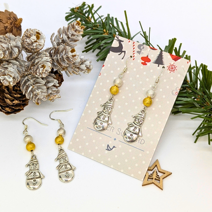 Very Merry Snowmen Earrings by Silver Sunbird—festive, handmade sterling silver earrings with gold bead accents, presented on a decorative card and artfully arranged with pine branches, pine cones, and a wooden star.