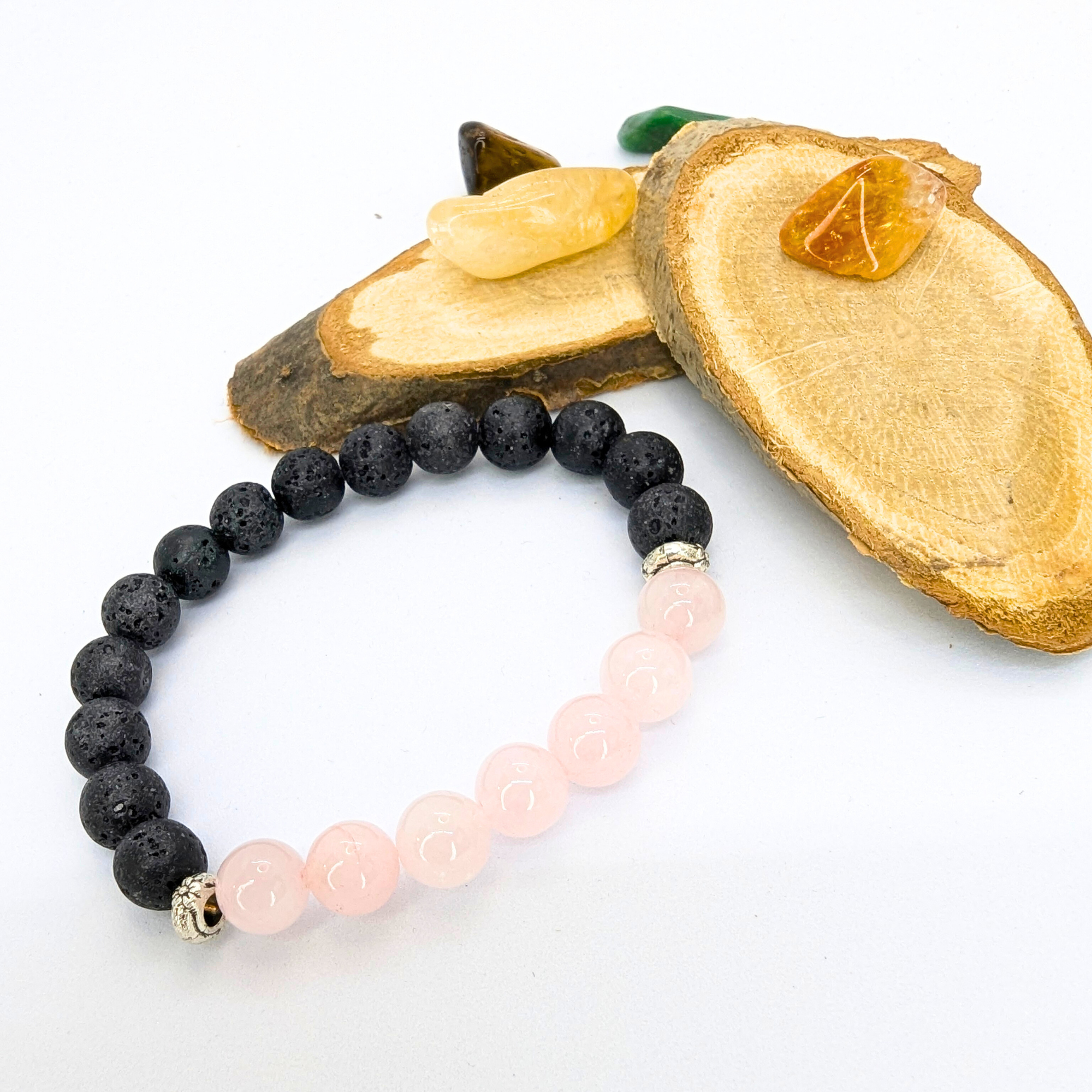 A Silver Sunbird Rose Quartz and Lava Stone bracelet featuring black and pink beads is elegantly displayed on two wooden slices, accompanied by small polished stones in shades of brown, green, and orange.