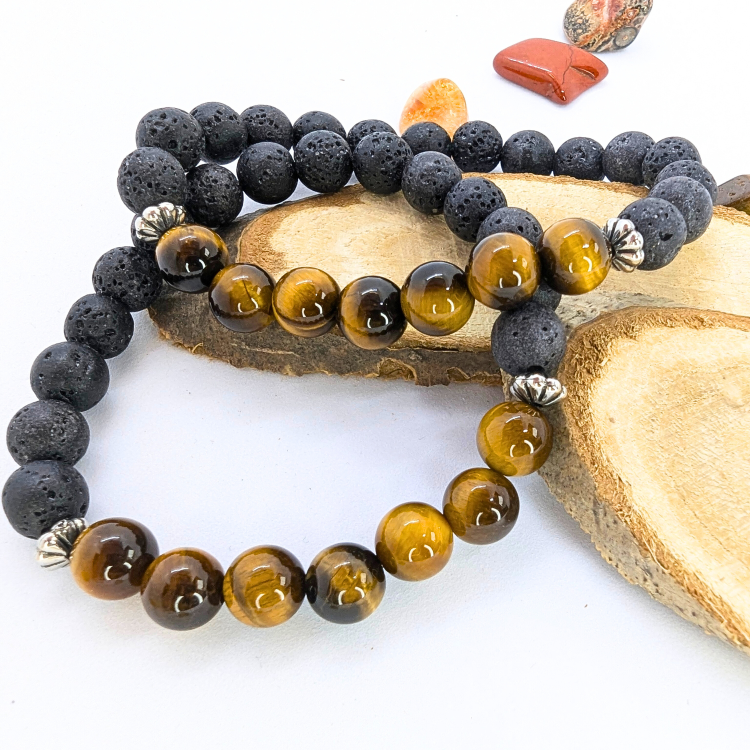 Two Silver Sunbird bracelets, part of the Tiger Eye and Lava Stone collection, rest on wood slices. One bracelet highlights lustrous Tiger&