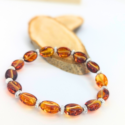 A Silver Sunbird Coffee Bean Bracelet with glossy, amber-colored oval beads and silver spacers is displayed on wood, its warm shine highlighted by a softly blurred backdrop—perfect for coffee lovers.