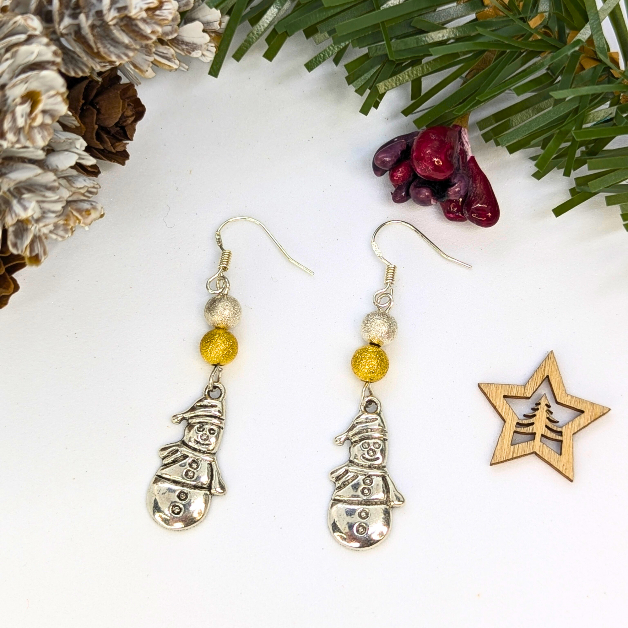The Very Merry Snowmen Earrings by Silver Sunbird are festive handmade pieces featuring silver charms with gold and white beads, shown on a white surface with pine cones, greenery, red berries, and a wooden star accent.