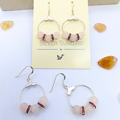 A pair of Rose Quartz Statement Earrings from Silver Sunbird displayed on a card labeled &quot;Silver Sunbird.&quot; The earrings feature circular silver hoops adorned with pink gemstones and small red rhinestone spacers. Four additional gemstones are scattered on a white surface around the card.