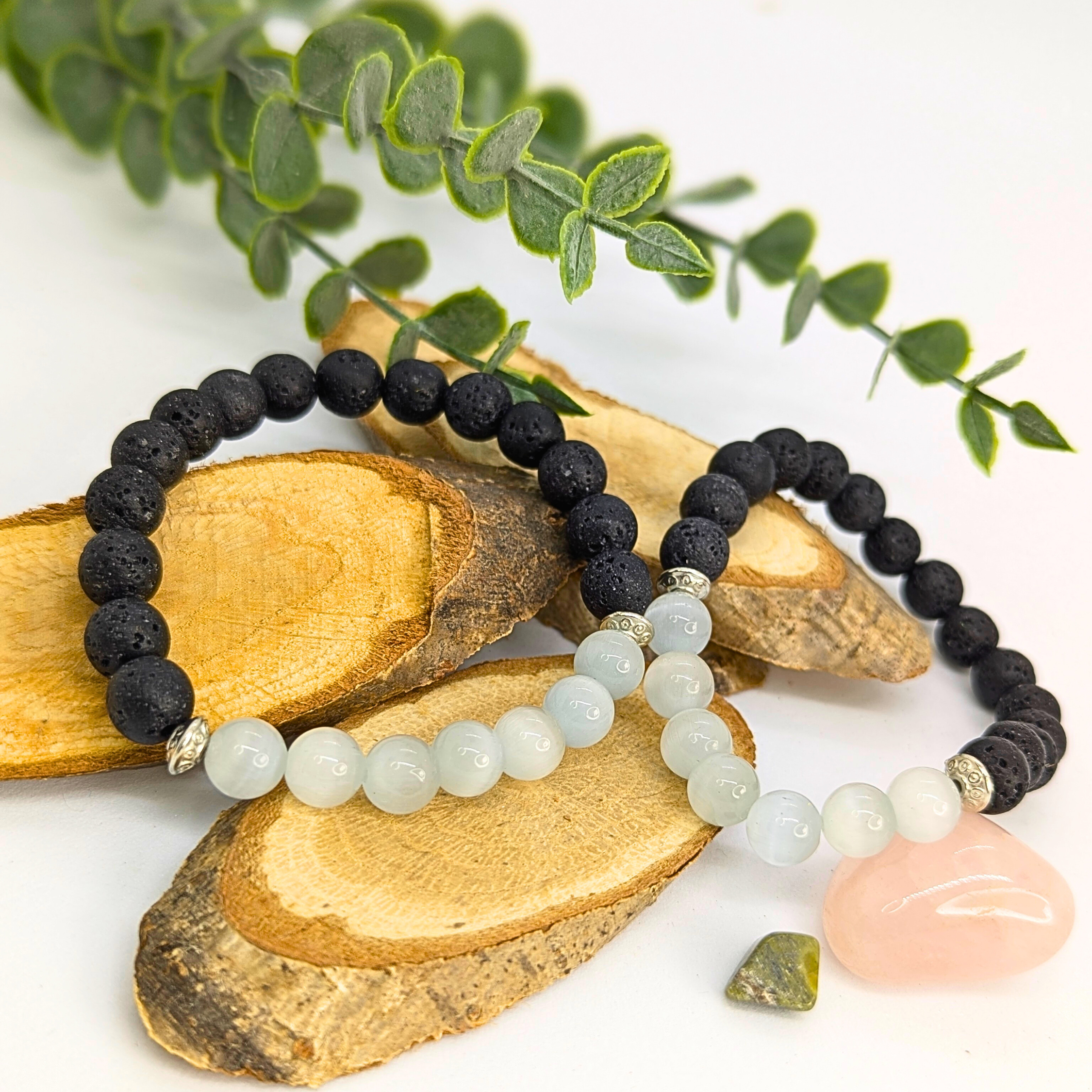 Silver Sunbird’s Moonstone and Lava Bracelets—one with black lava stones, the other with light blue moonstone beads—are displayed on wood slices, accented by a pink stone, green stone, and decorative green leaves.