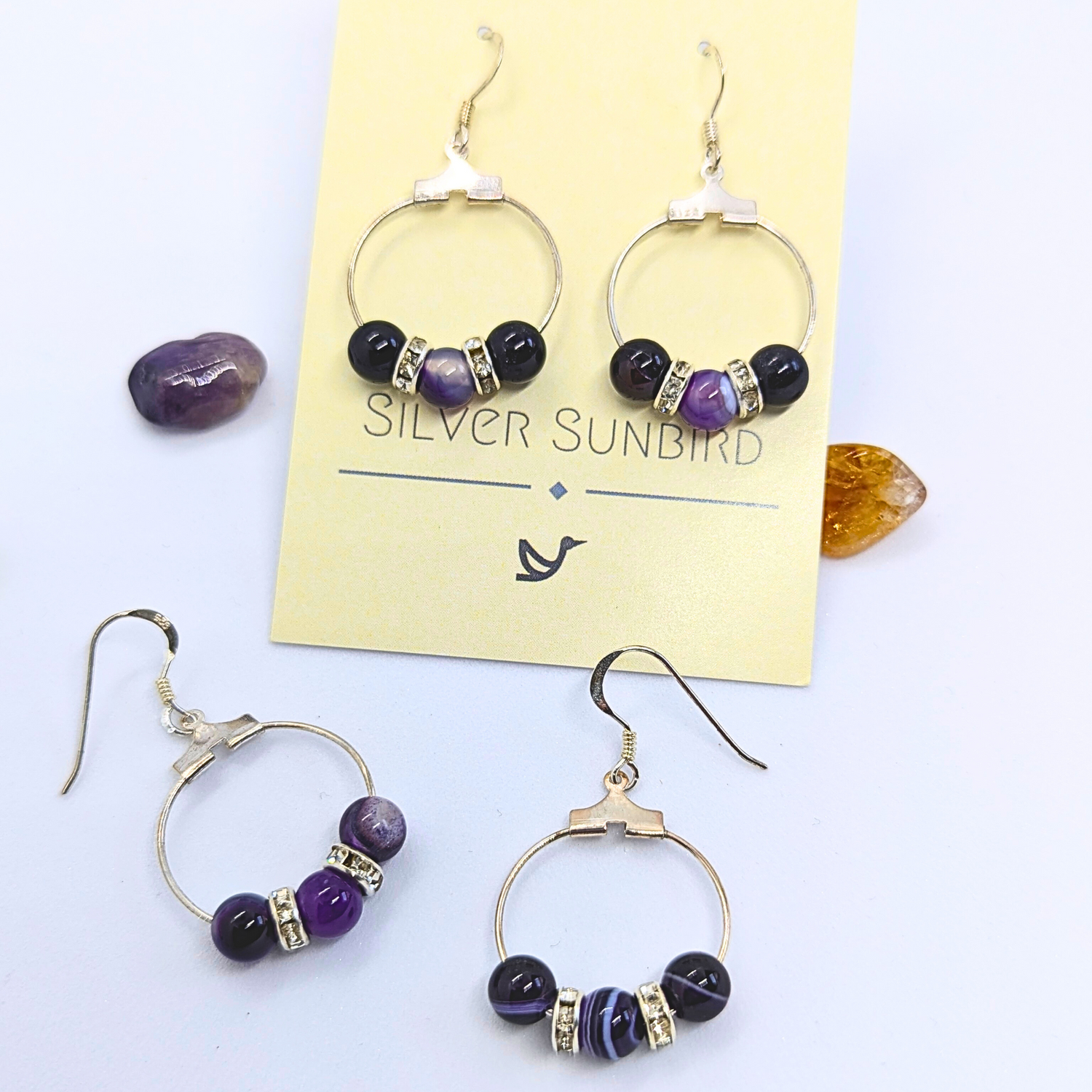 A pair of stylish Amethyst Statement Large Earrings lay on a card labelled &quot;Silver Sunbird.&quot; The sterling silver earrings are circular with purple amethyst round gemstones, adorned with small silver rhinestone spacers. Beside the card are two loose matching earrings and three natural gemstones in shades of purple and amber.