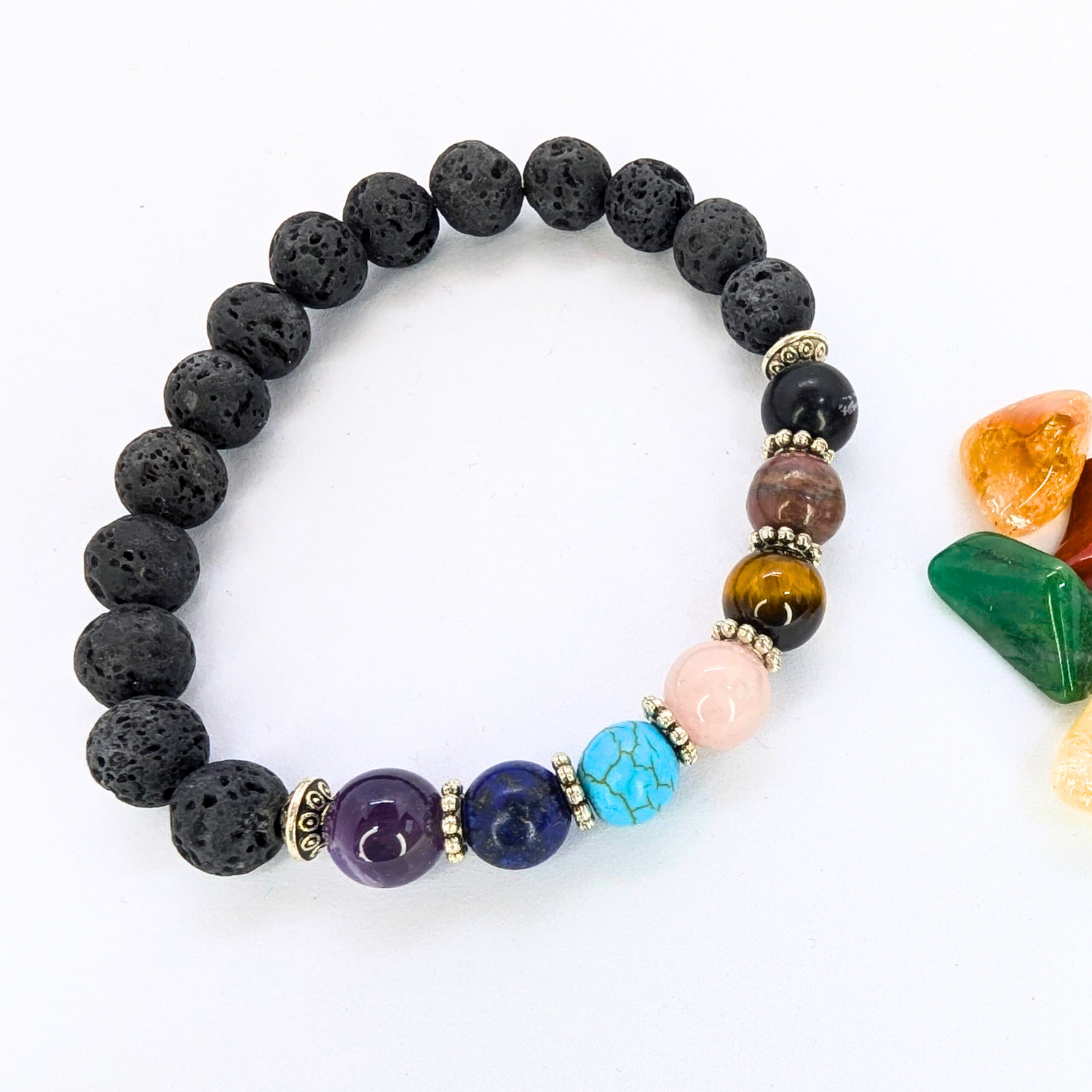 The Chakra Bracelet by Silver Sunbird, featuring black lava stones and seven colorful gemstones, is shown on a white surface beside small polished green, orange, and yellow stones.