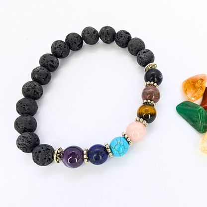 The Chakra Bracelet by Silver Sunbird, featuring black lava stones and seven colorful gemstones, is shown on a white surface beside small polished green, orange, and yellow stones.