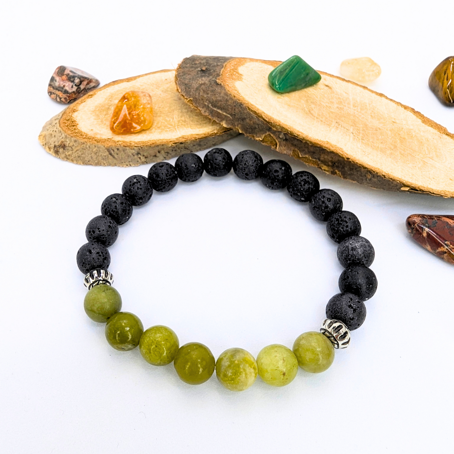 The Silver Sunbird Peridot Bracelets, featuring alternating black and green beads with silver accents, are elegantly displayed on a white background surrounded by slices of wood and small colorful stones.