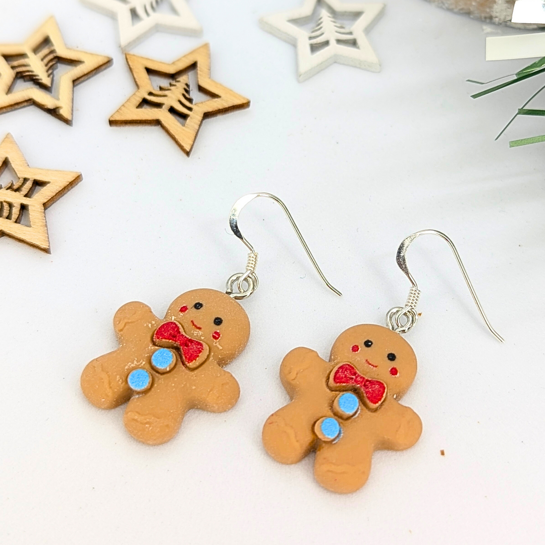 The Cheerful Gingerbread Man Earrings by Silver Sunbird, featuring red bows and blue buttons, are displayed on a white surface. Wooden star decorations are scattered around to enhance the festive atmosphere.