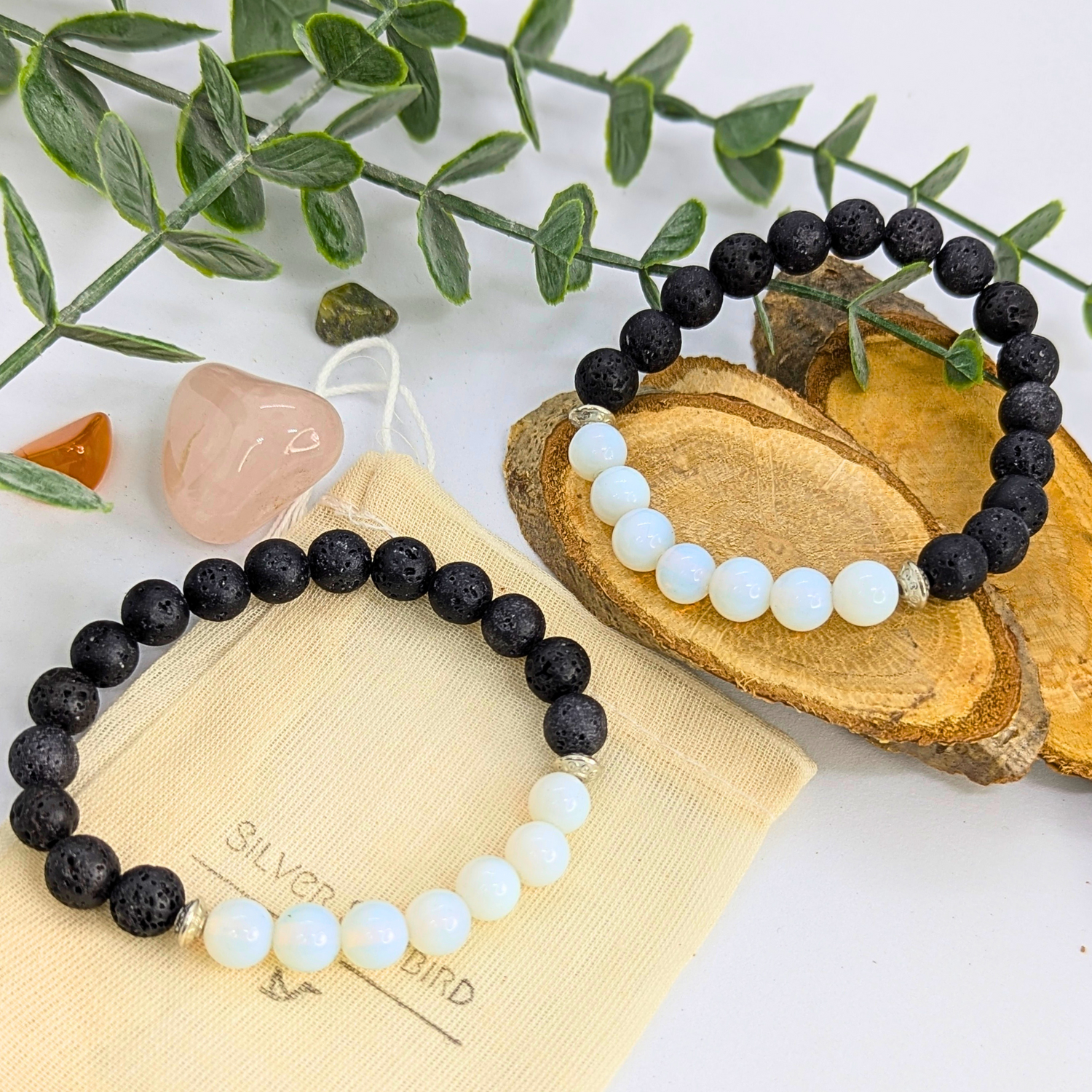 The Silver Sunbird Opal and Lava Stone Bracelets, featuring alternating black and white stones, are displayed on wooden slices with a beige pouch, accented by crystals and green leaves.