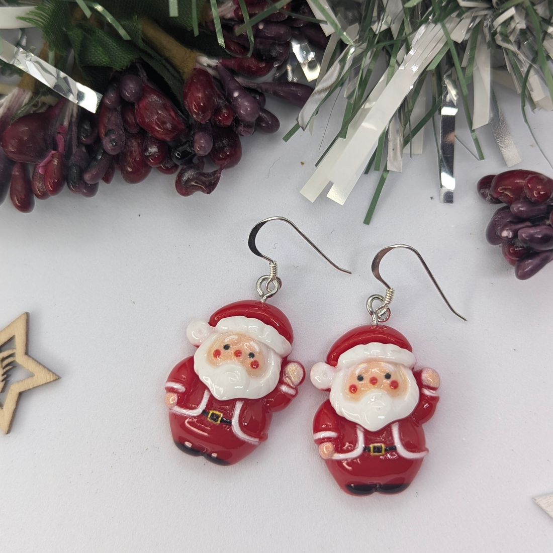 The Jolly Santa Earrings by Silver Sunbird rest on a white surface, surrounded by pine branches and berries, epitomizing the charm of Christmas accessories.