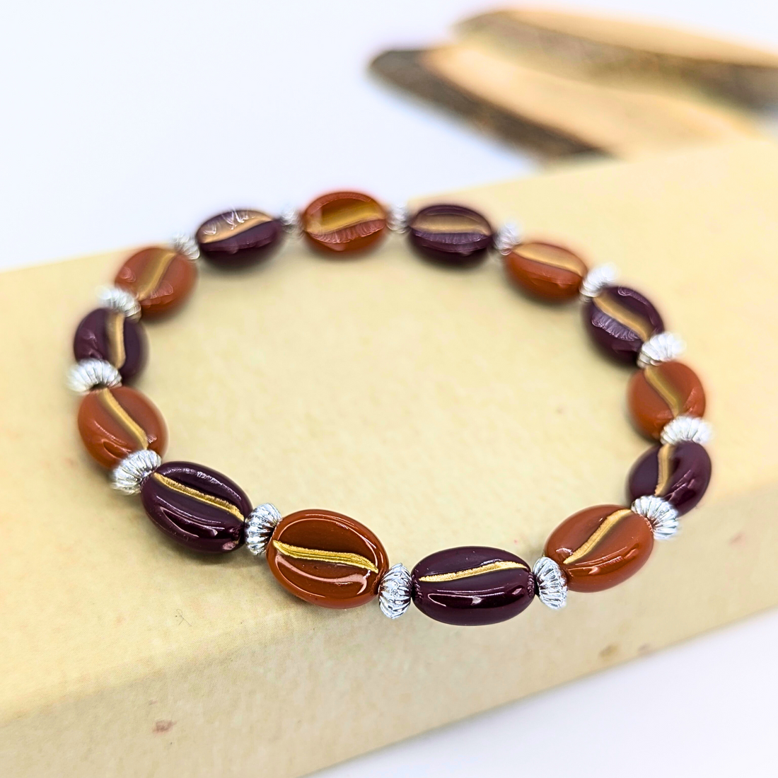 The Silver Sunbird Coffee Bean Bracelet showcases exquisite oval beads in brown and dark maroon with central gold stripes, interspaced by small silver-toned accents. Ideal for coffee lovers, it&