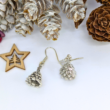 Silver Sunbird’s 3D Christmas Tree Earrings in sterling silver rest on a white surface, surrounded by frosted and brown pinecones, plus a wooden star ornament with a Christmas tree charm cutout.