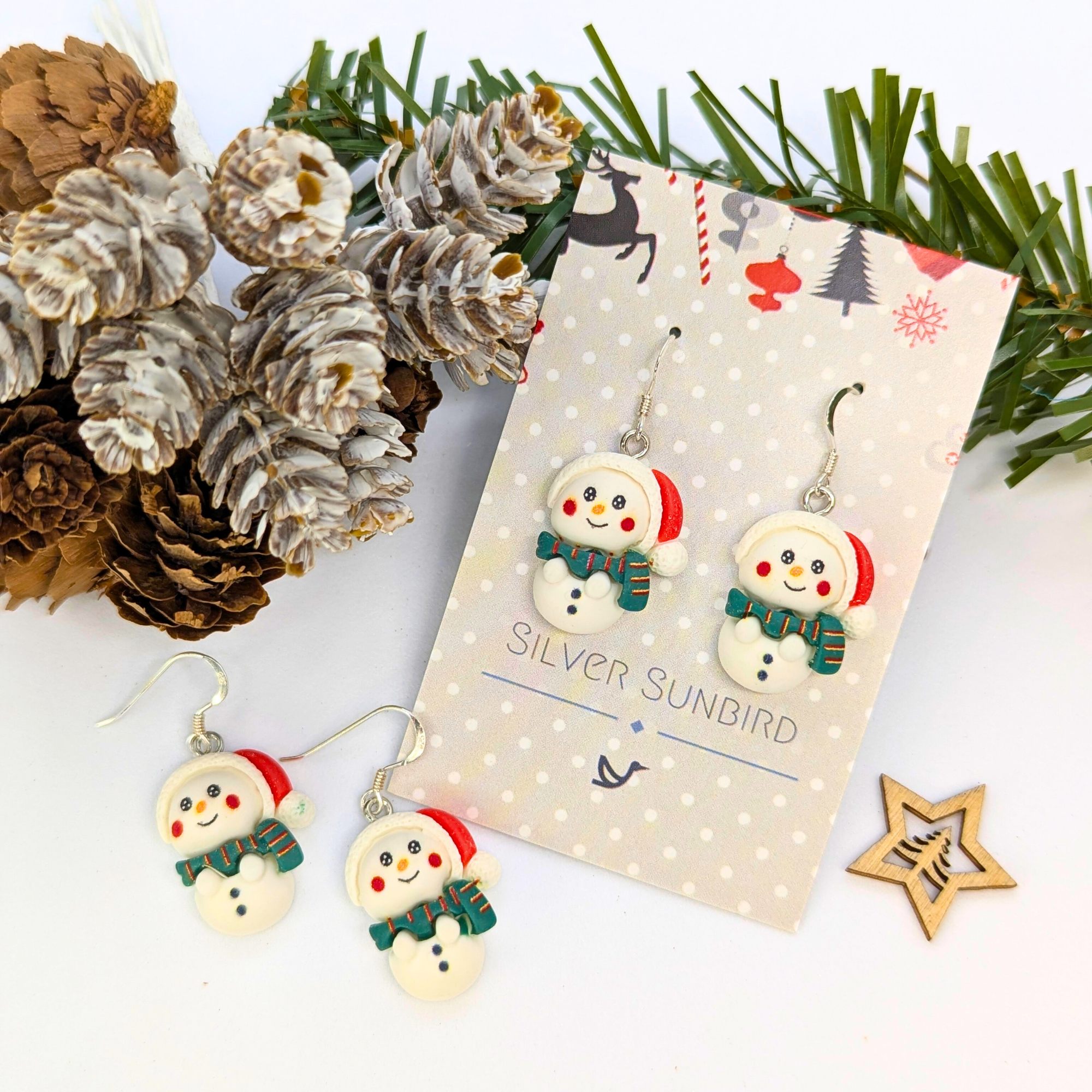 Three pairs of Bright Snowmen Earrings by Silver Sunbird, featuring scarves and red earmuffs, are displayed on a branded card among pinecones, greenery, and a wooden star—ideal for festive flair in your Christmas jewelry collection.