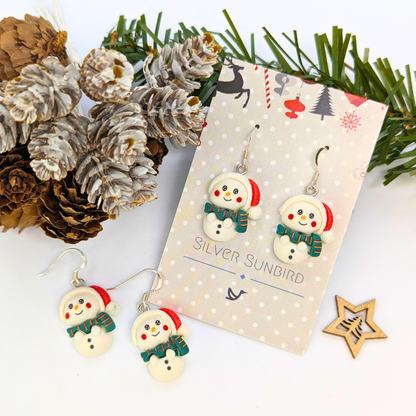 Three pairs of Bright Snowmen Earrings by Silver Sunbird, featuring scarves and red earmuffs, are displayed on a branded card among pinecones, greenery, and a wooden star—ideal for festive flair in your Christmas jewelry collection.