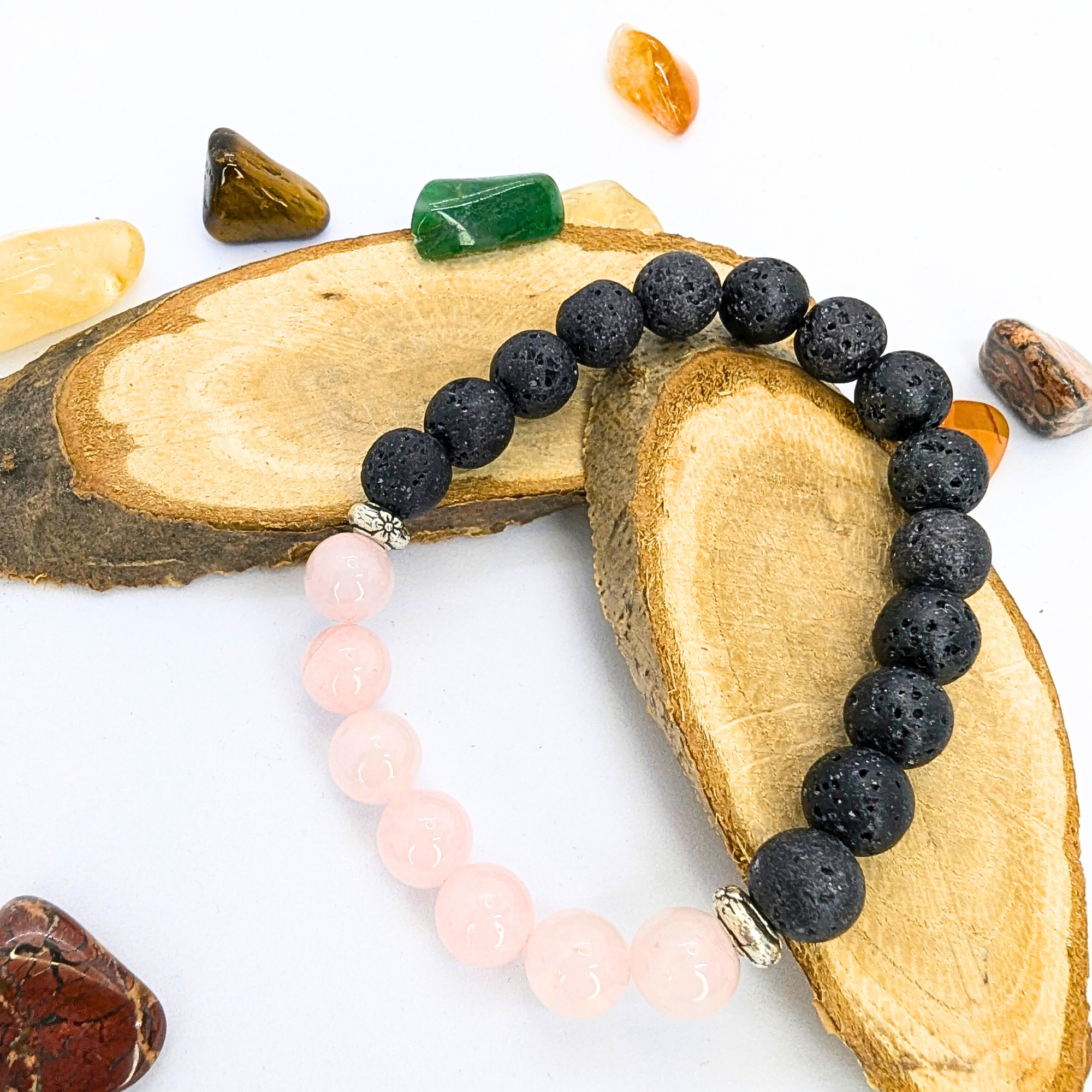 A Silver Sunbird Rose Quartz and Lava Stone Bracelet with pink rose quartz beads featuring silver spacers and textured black lava beads rests on two wooden slices. Surrounding the bracelet are scattered various small, colorful stones against a white background.