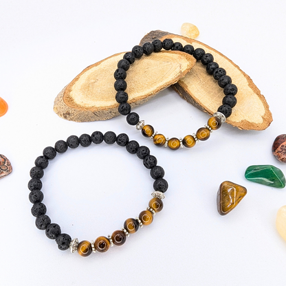 The Silver Sunbird Tiger Eye and Lava Stone Bracelets, featuring black and brown stones with silver accents, are displayed on wooden slices surrounded by polished orange, green, and reddish-brown gemstones on a light-colored surface.