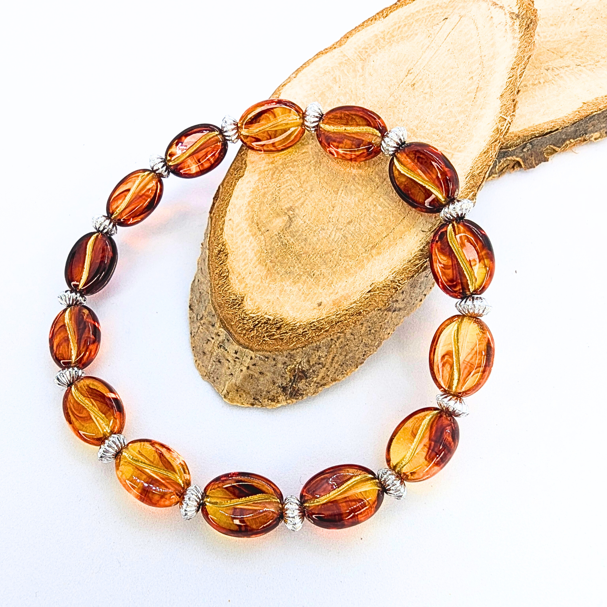 The Silver Sunbird Coffee Bean Bracelet showcases elegant amber oval beads and silver spacers on a natural wood slice against a white backdrop, making it a perfect stylish accessory for coffee lovers.