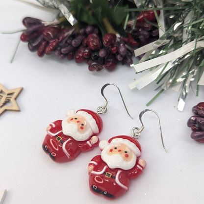 The Jolly Santa Earrings by Silver Sunbird are the perfect Christmas accessories, featuring jolly Santa figures in red suits and white beards. They rest on a white surface adorned with festive greenery and purple berries, encapsulating the charm of holiday jewelry.