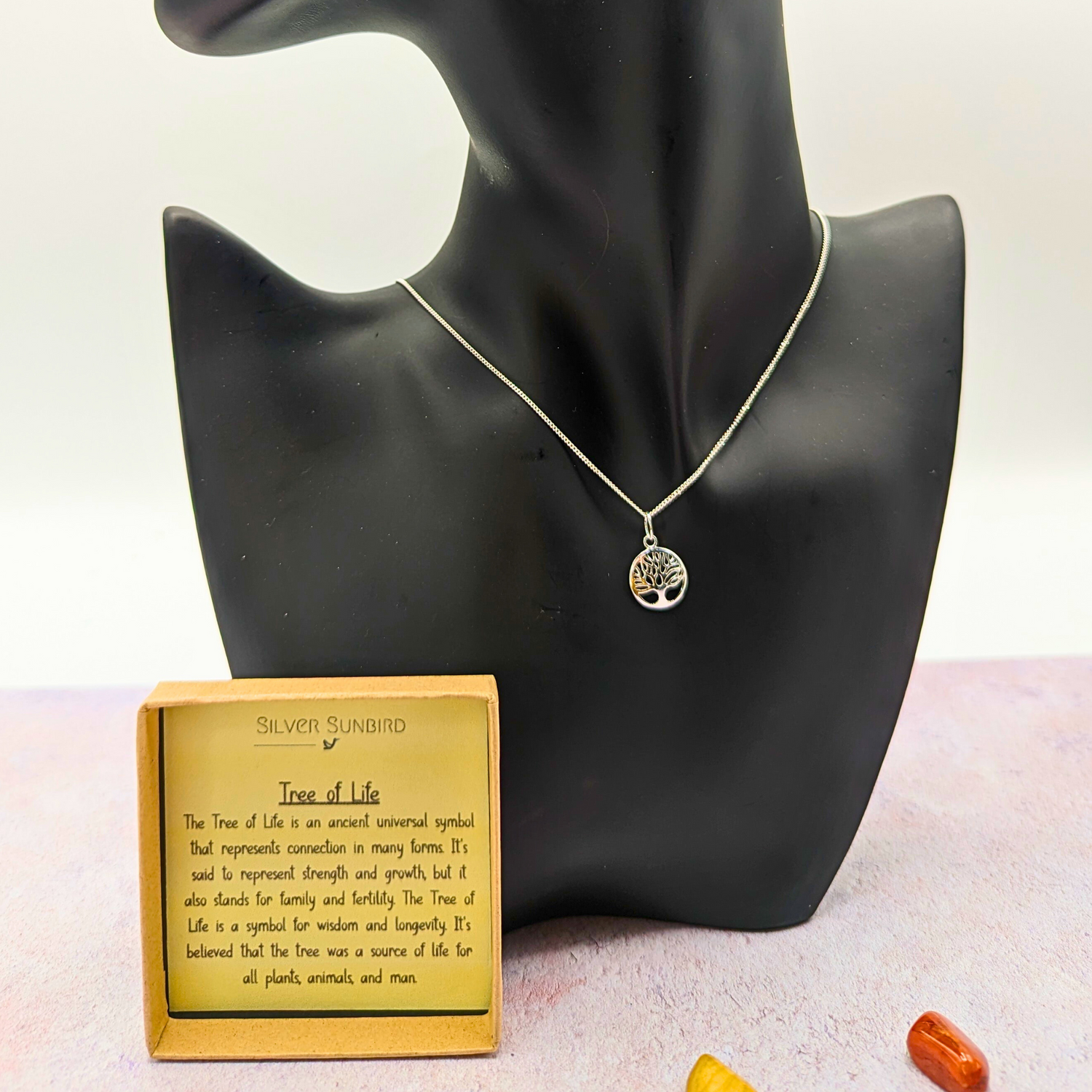 The Silver Sunbird Tree of Life Sterling Silver Necklace is showcased on a black bust, accompanied by an informative card. Set on a bright surface with colorful stones, it&