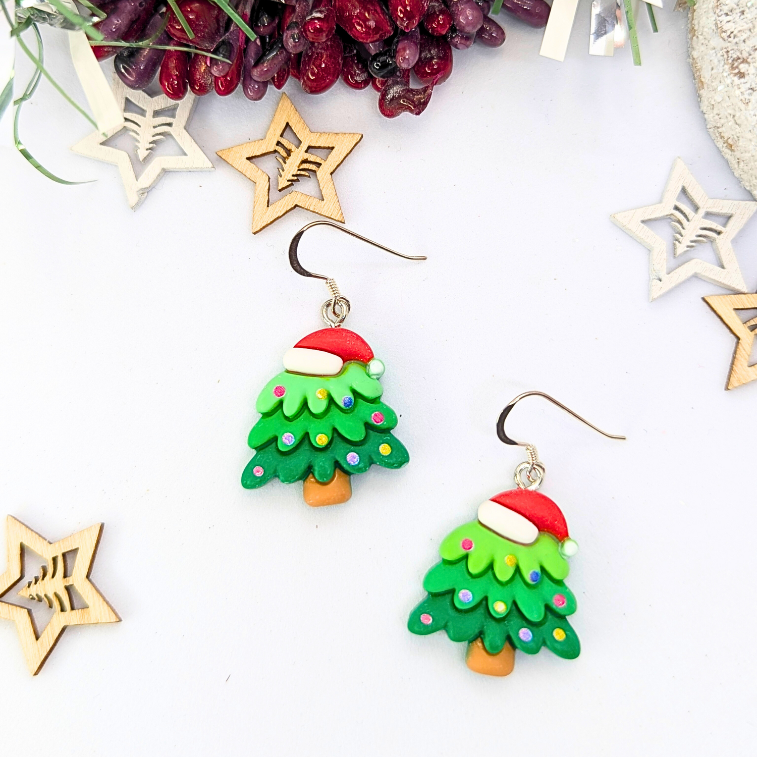 The Christmas Tree Earrings by Silver Sunbird feature festive designs with Santa hats and colorful dots, elegantly showcased against a white background. Accompanied by wooden star ornaments and vibrant red berries, these earrings are the perfect holiday accessory.