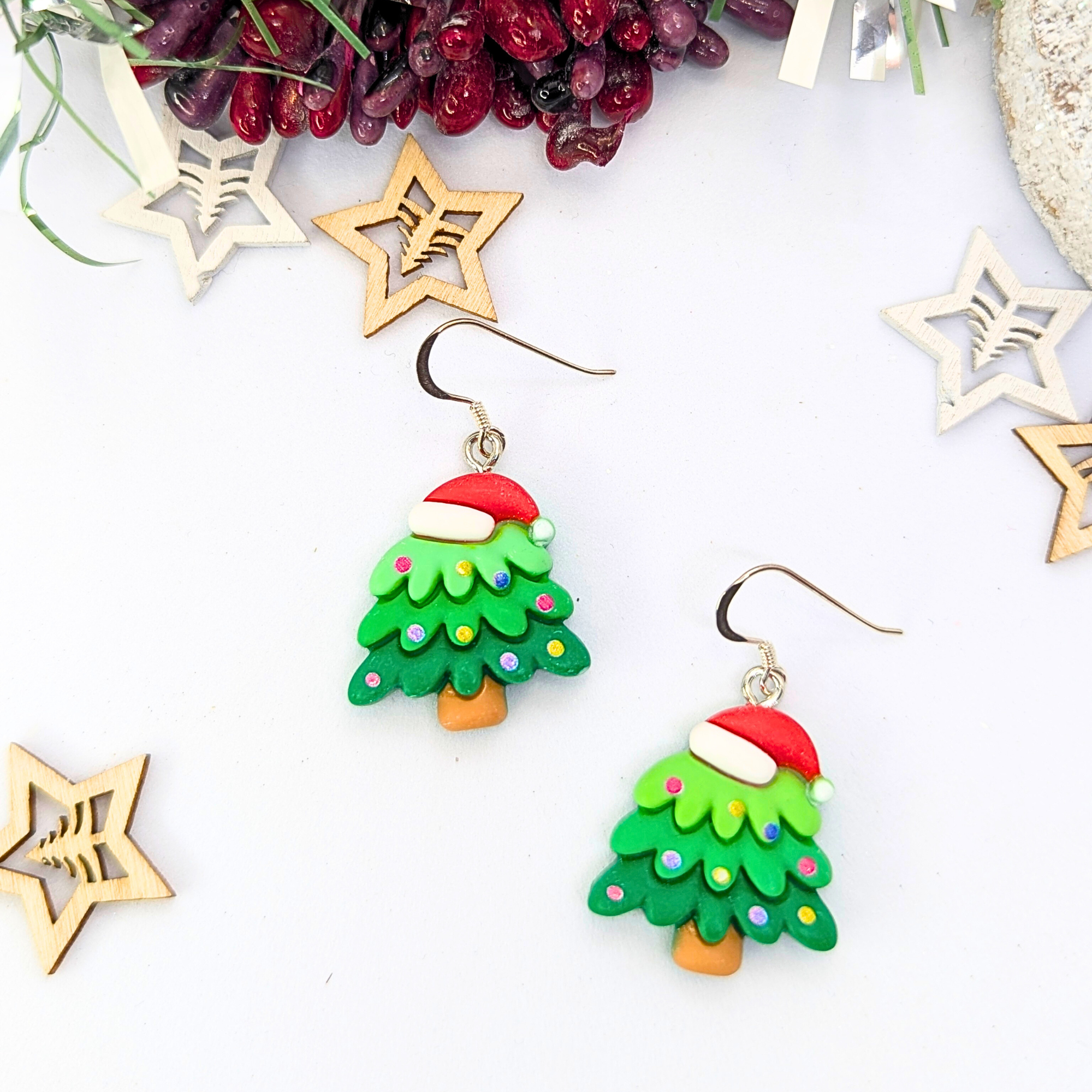 The Christmas Tree Earrings by Silver Sunbird feature festive designs with Santa hats and colorful dots, elegantly showcased against a white background. Accompanied by wooden star ornaments and vibrant red berries, these earrings are the perfect holiday accessory.