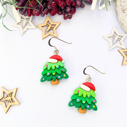 The Christmas Tree Earrings by Silver Sunbird feature festive designs with Santa hats and colorful dots, elegantly showcased against a white background. Accompanied by wooden star ornaments and vibrant red berries, these earrings are the perfect holiday accessory.