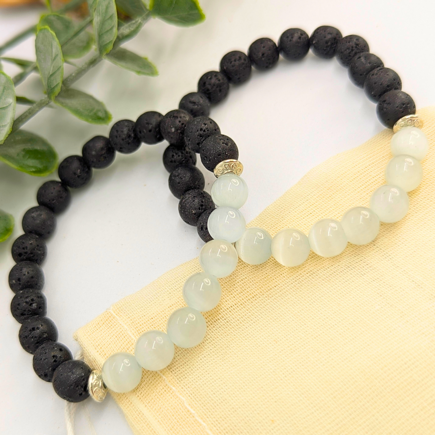 The Silver Sunbird Moonstone and Lava Bracelets—two handcrafted pieces with alternating black and white round beads—are displayed on a light fabric pouch, accented by green leaves in the background.