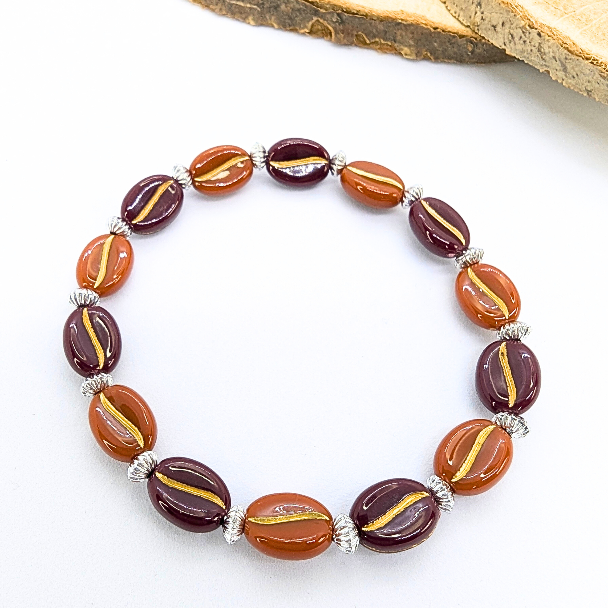 The Silver Sunbird Coffee Bean Bracelet features alternating brown and maroon oval beads with gold lines, separated by silver spacers. It&