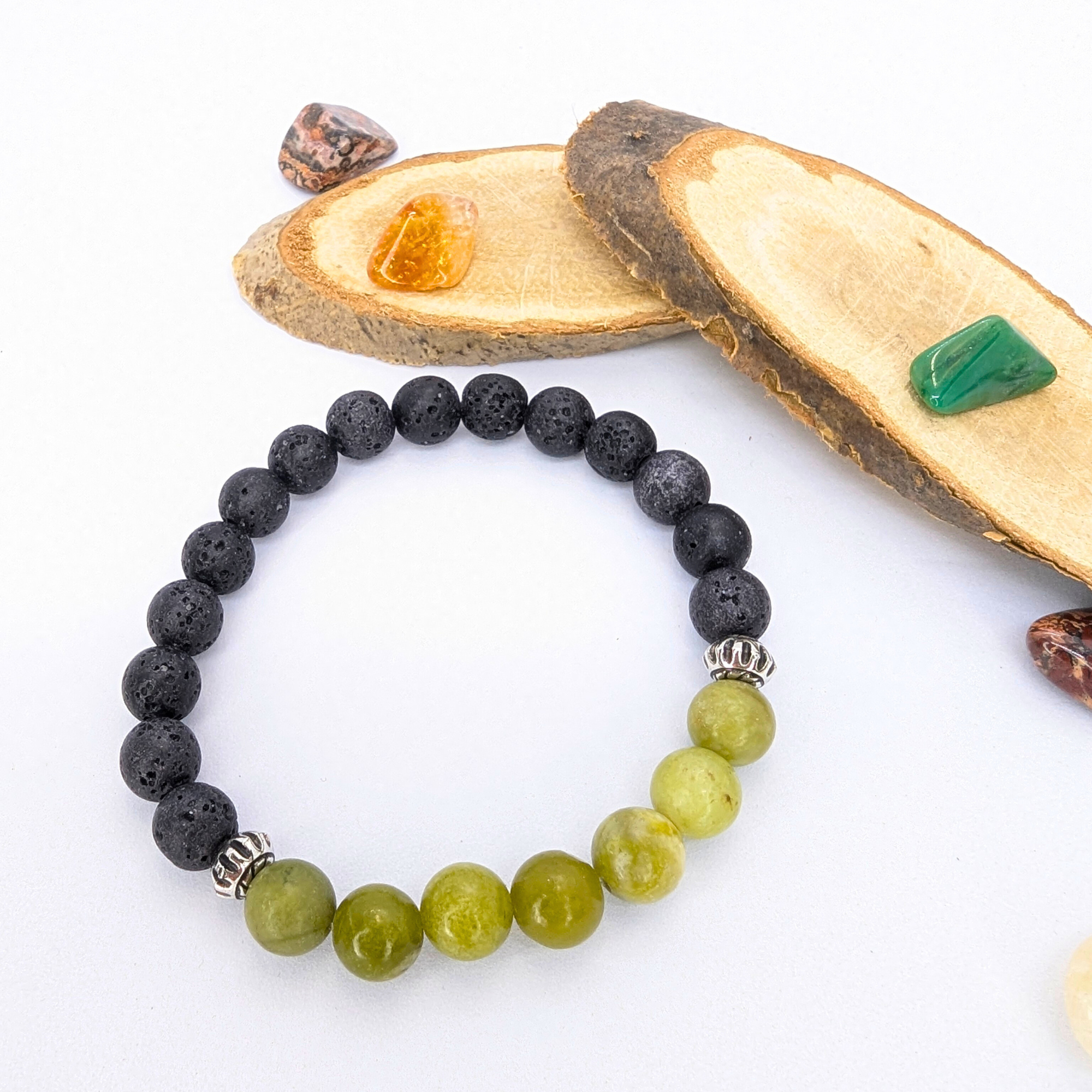 The Silver Sunbird Peridot Bracelets, featuring black and green beads, are displayed on a white surface alongside smooth wooden pieces and vibrant gemstone jewelry in shades of orange, brown, and green.