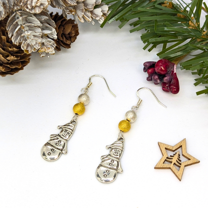 Very Merry Snowmen Earrings by Silver Sunbird: festive handmade earrings featuring silver snowman charms with gold and silver beads, displayed on white with pinecones, pine branches, red berries, and a wooden star ornament.