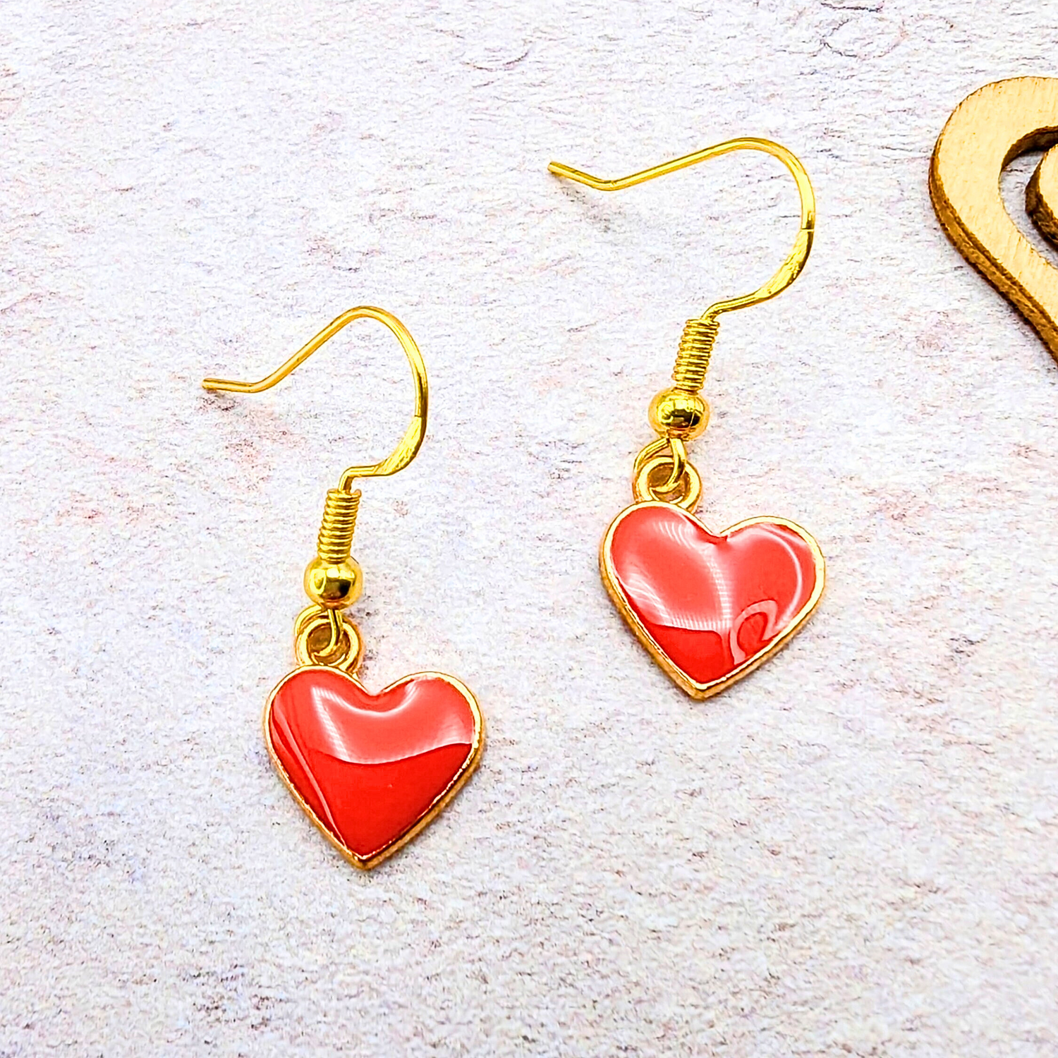 The Alluring Red Heart Earrings by Silver Sunbird feature vibrant red centers and hypoallergenic gold hooks, elegantly displayed on a textured surface beside a wooden cutout, offering a charming accessory that is both stylish and gentle on the skin.