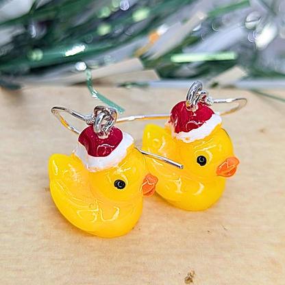 A close-up image showcases the Festive Duck Earrings by Silver Sunbird, adorned with yellow rubber ducks wearing red and white Santa hats. The blurred background of green and white exudes holiday cheer, while these handcrafted accessories rest on a light wooden surface, ideal for adding a playful touch to any seasonal outfit.
