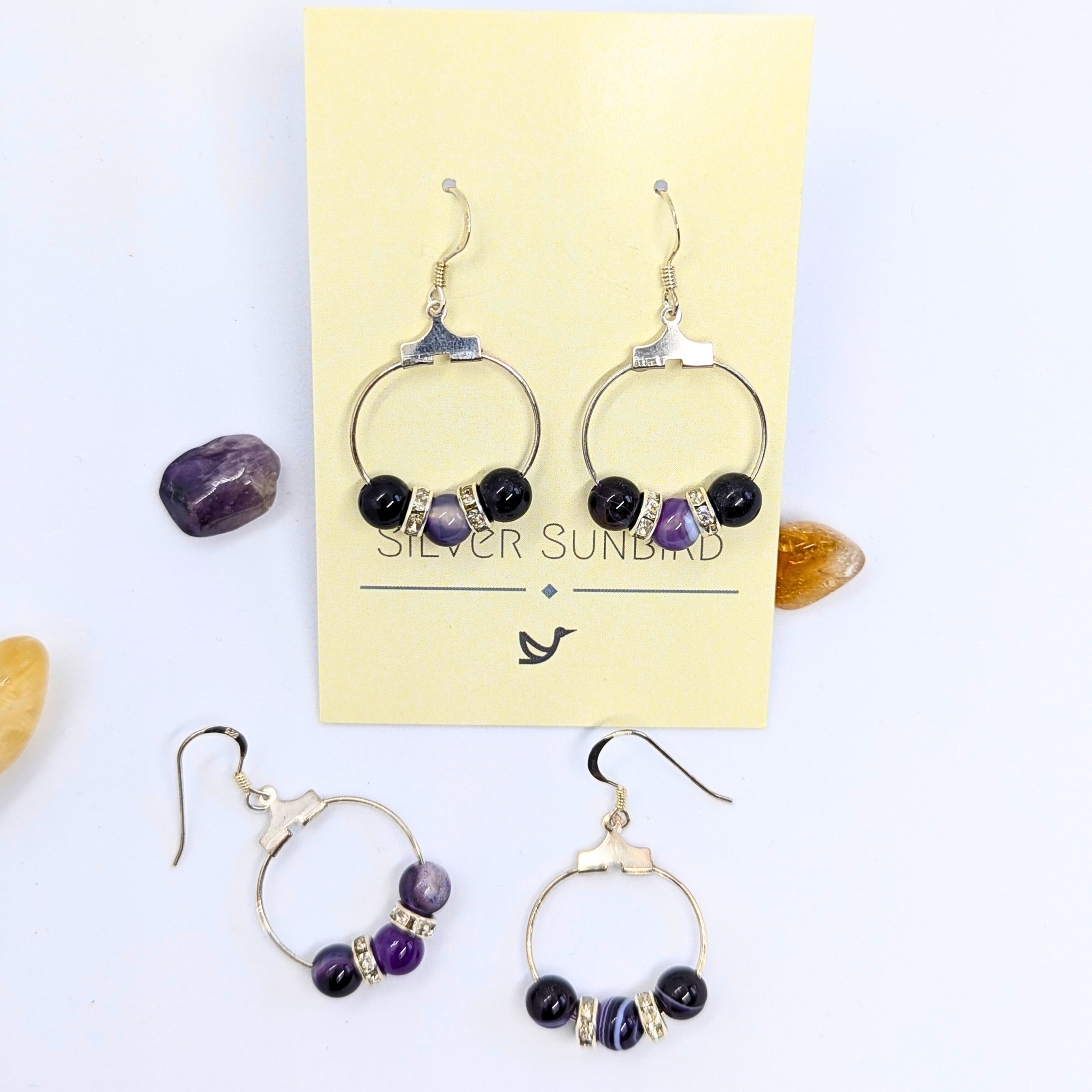 A set of Amethyst Statement Large Earrings by Silver Sunbird displayed on a light yellow card. The earrings, crafted from 925 sterling silver, feature circular hoops adorned with dark purple amethyst. Surrounding the card are small polished stones in amethyst and citrine shades, adding a touch of natural gemstones.