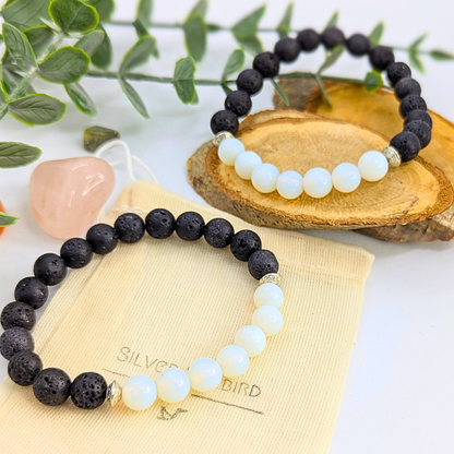 The Silver Sunbird Opal and Lava Stone Bracelets, featuring alternating black lava beads and white opal, are displayed on a cream pouch atop a wooden tray, surrounded by green leaves and a pink crystal.