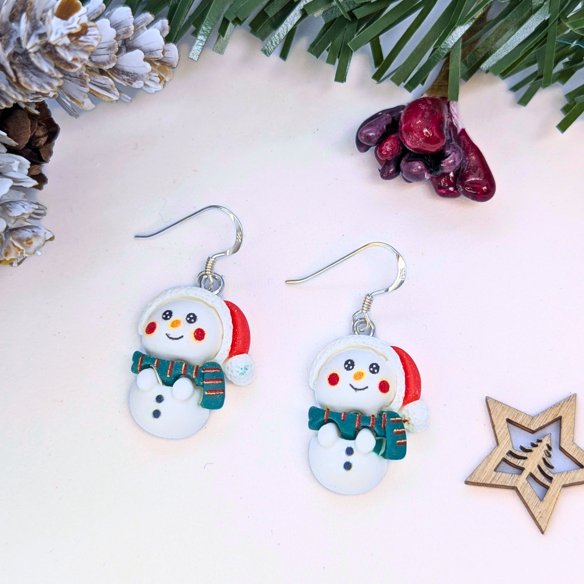 Two Bright Snowmen Earrings by Silver Sunbird, shaped like smiling snowmen with red Santa hats and green-striped scarves, make festive Christmas jewelry. Shown on a white surface with evergreen branches, pinecones, berries, and a wooden star.