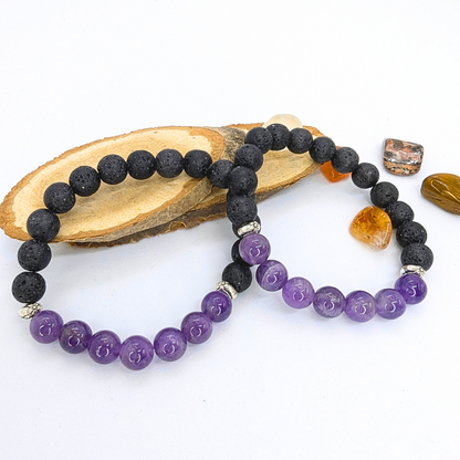 Two stretch bracelets made with black lava stones and purple amethyst beads are displayed on a wooden slice, surrounded by small polished stones in orange and white. The amethyst beads are smooth and glossy, while the lava stones have a textured, porous appearance.