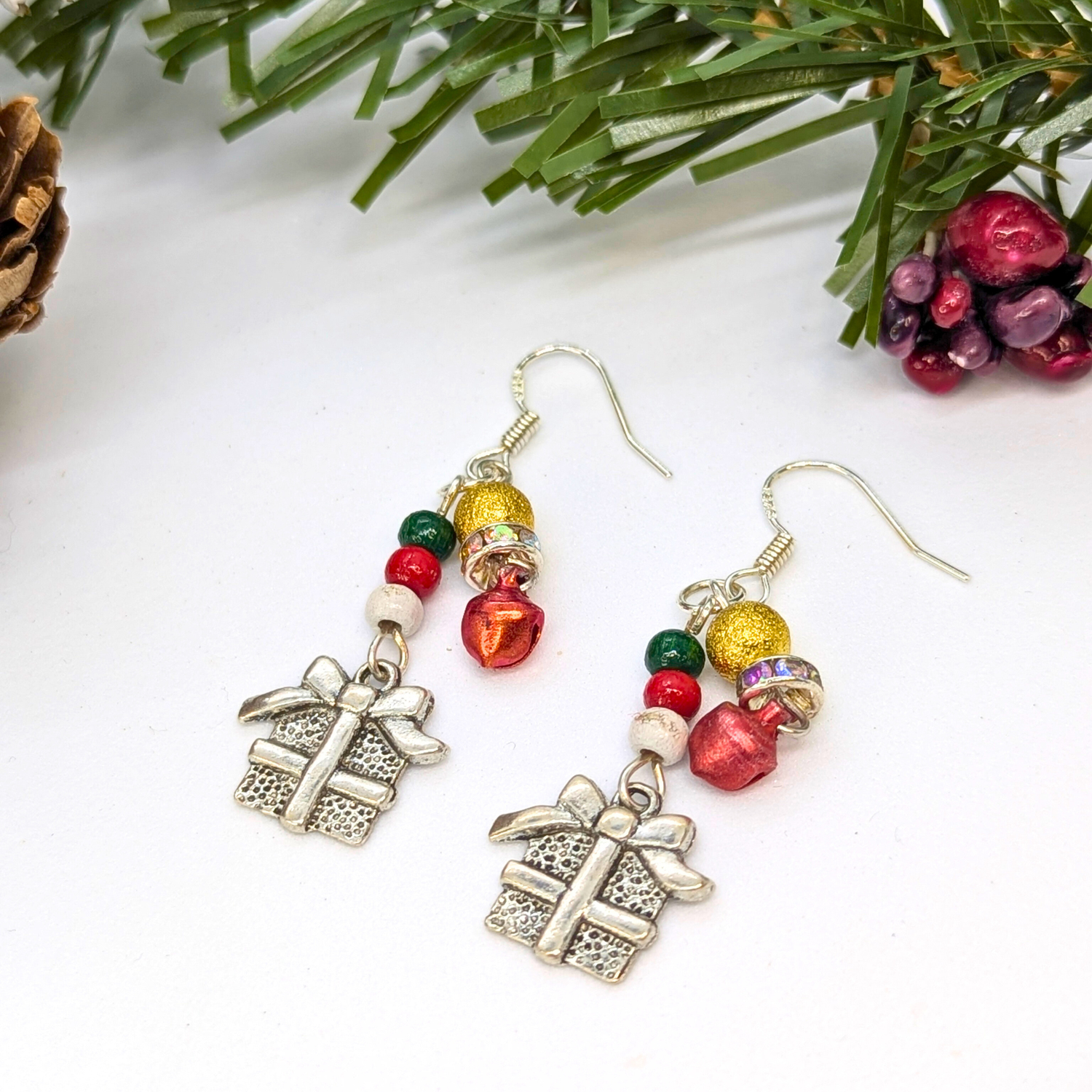 Silver Sunbird Gift Box Dangle Earrings feature red and gold beads, jingle bells, and festive gift charms. Accented with sterling silver hooks, they rest on a white surface with pine and berries for a holiday touch.