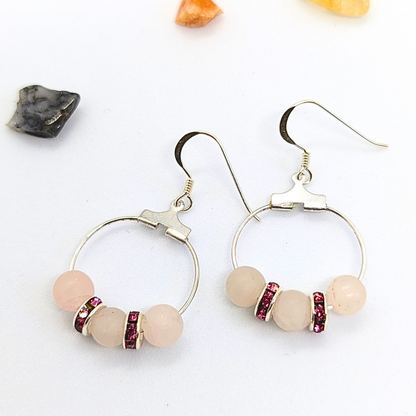 The Rose Quartz Statement Earrings by Silver Sunbird feature a pair of sterling silver hoops, each adorned with three round rose quartz beads, separated by two pink rhinestone spacers. These hypoallergenic earrings are displayed on a white surface decorated with small decorative stones.