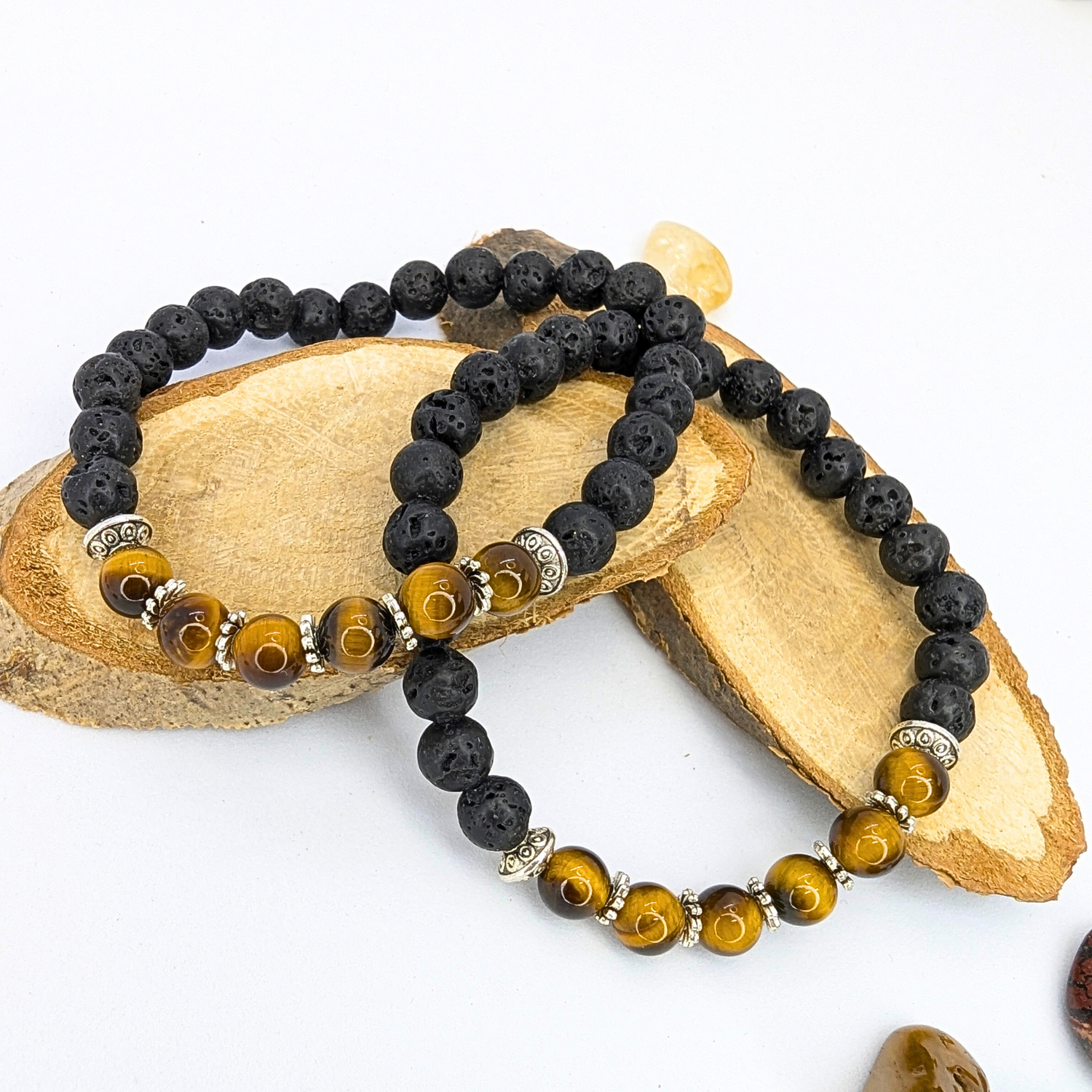 Two Tiger Eye and Lava Stone Bracelets by Silver Sunbird rest on wooden slices. Each bracelet features black lava stone beads, Tiger&