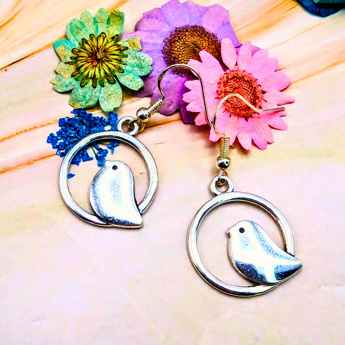 Silver Sunbird presents Beautiful Bird Earrings crafted for bird lovers, glistening within circular frames on a light wooden surface. Vibrant dried flowers in shades of pink, purple, green, and blue enhance the serene backdrop.