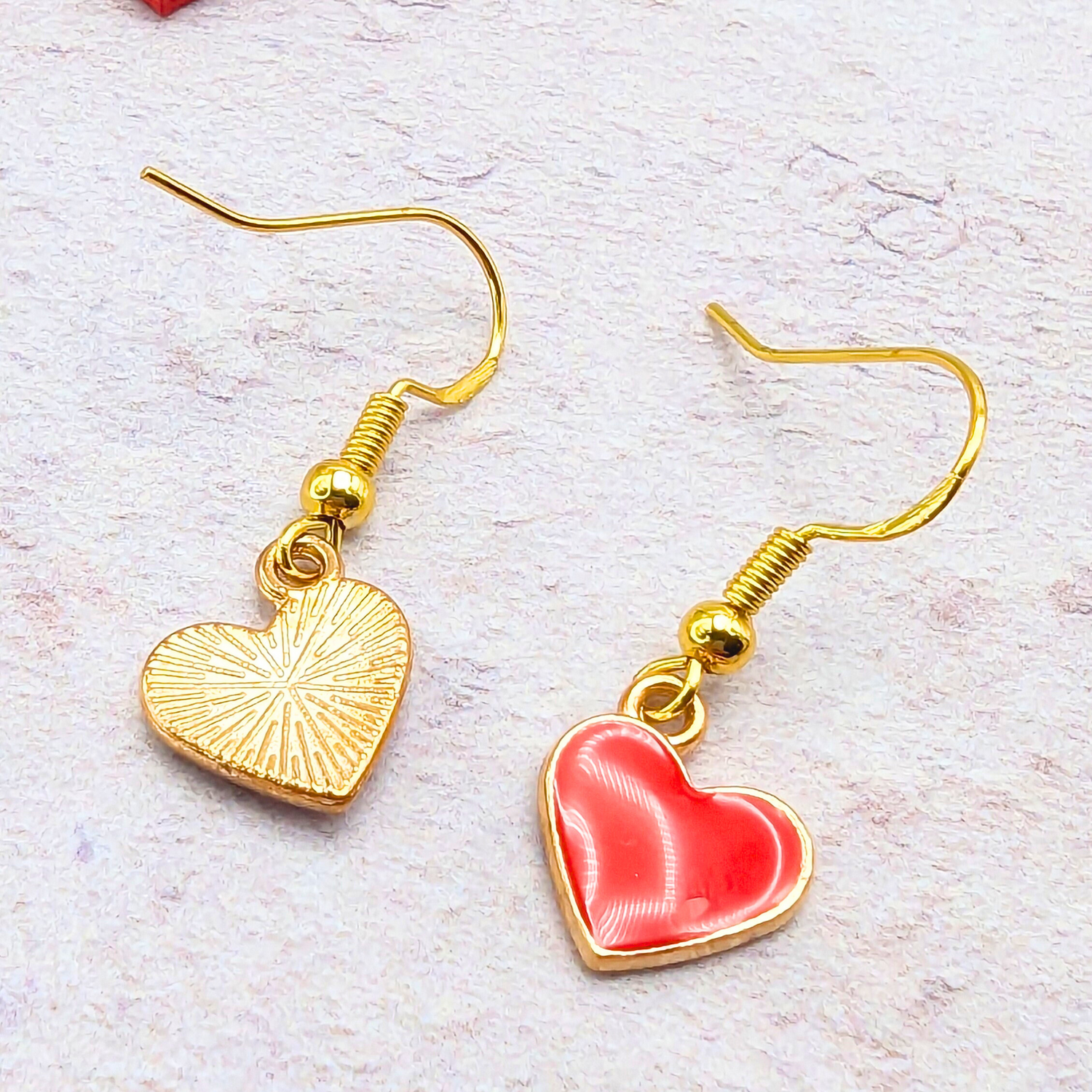 The Alluring Red Heart Earrings by Silver Sunbird feature two beautiful gold hook earrings with heart charms: one is textured in gold, and the other is embellished with red enamel outlined in gold. These hypoallergenic earrings rest gracefully on a light textured background, ideal for sensitive ears.