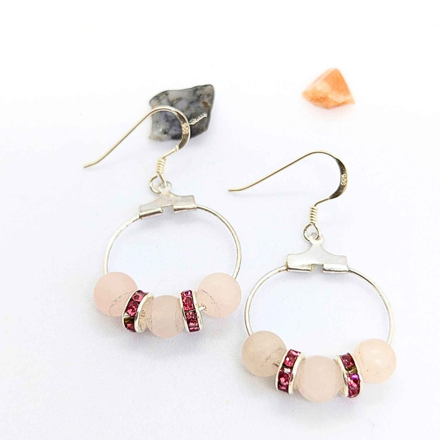 The elegant Rose Quartz Statement Earrings by Silver Sunbird feature hypoallergenic dangle design with circular silver hoops adorned with rose quartz gemstones and accented by small red spacers. The sterling silver earrings are beautifully showcased on a white surface alongside two small decorative stones — one black and white, the other orange.