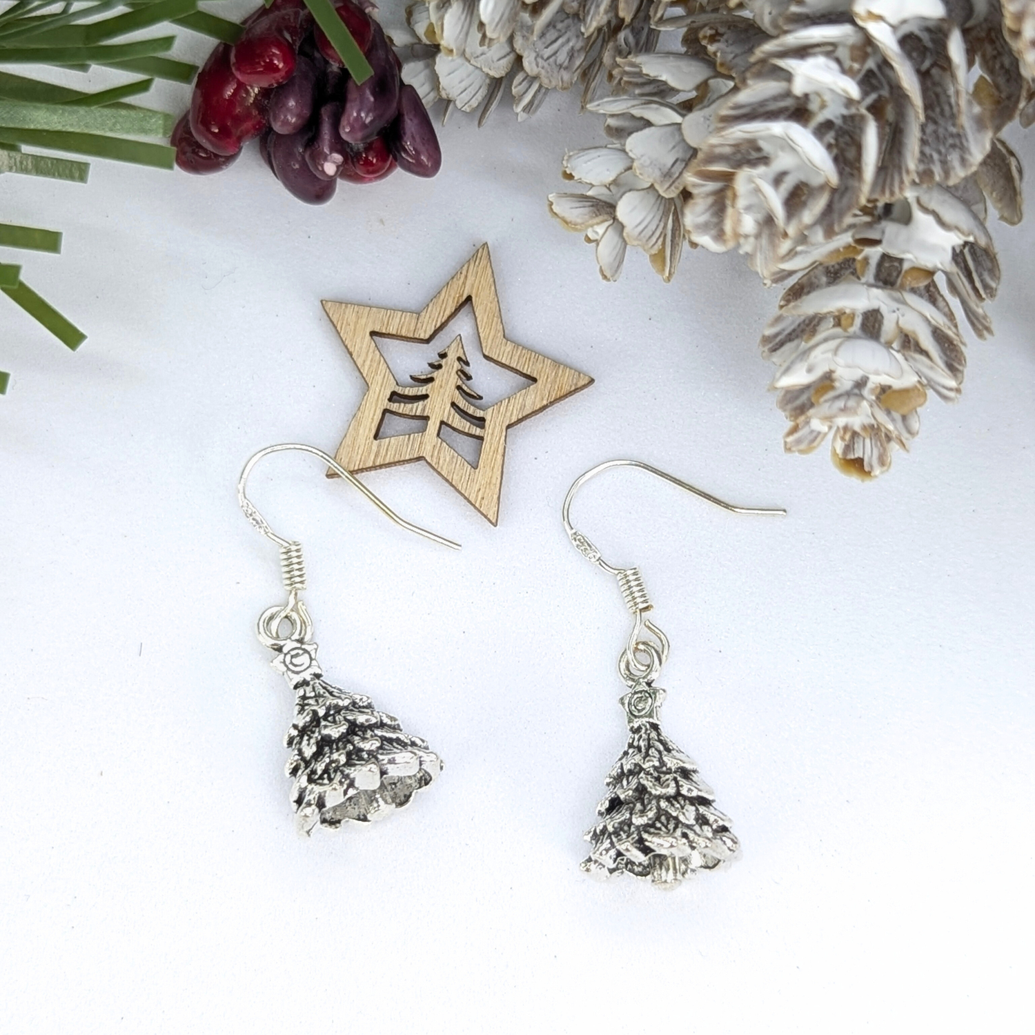 A pair of Silver Sunbird 3D Christmas Tree Earrings is displayed beside a wooden star ornament and frosted pinecones with red berries, all set on a white surface.