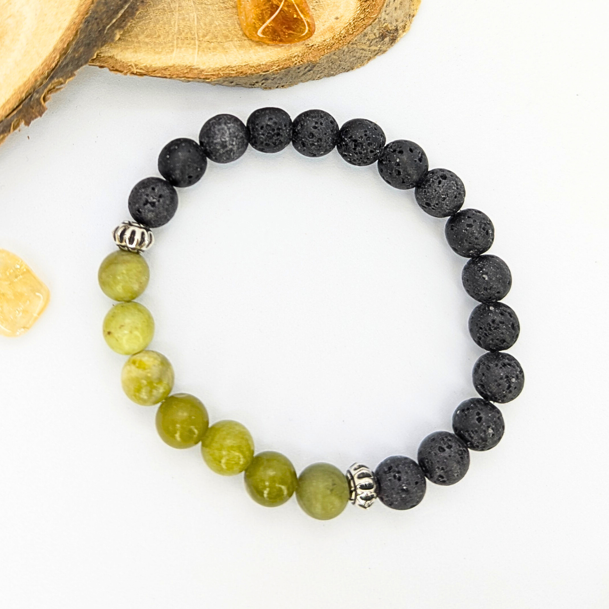 The Silver Sunbird Peridot Bracelet, featuring black and green stones with silver accents, is arranged in a circle on a white surface. Natural wooden slices and amber stones are visible in the background, highlighting exquisite gemstone jewelry.