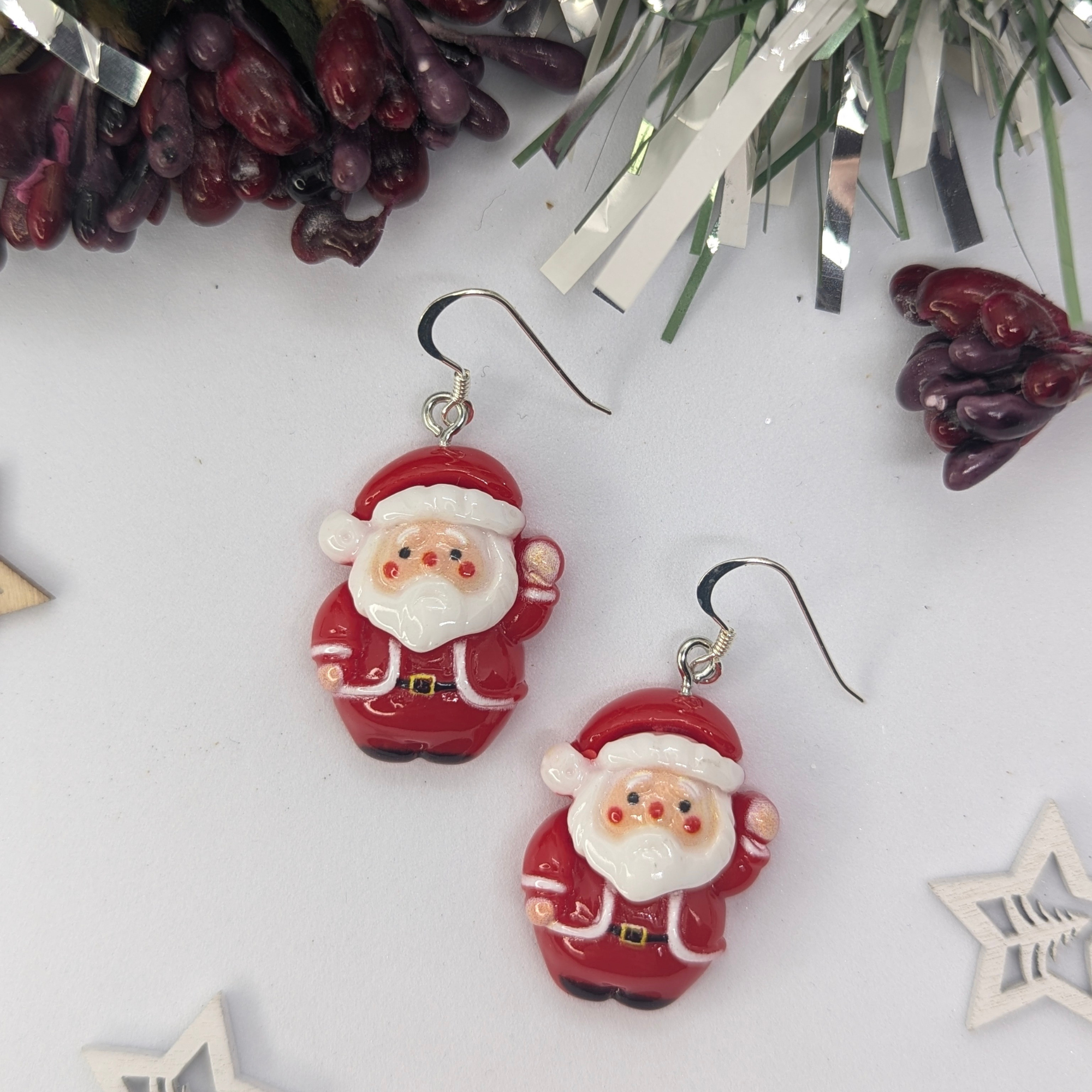 The Jolly Santa Earrings by Silver Sunbird dazzle with their cheerful design, featuring rosy-cheeked, jolly figures dressed as Santa Claus. Set against a white background adorned with festive holiday decorations and star-shaped embellishments, these Christmas accessories perfectly embody the spirit of the season.