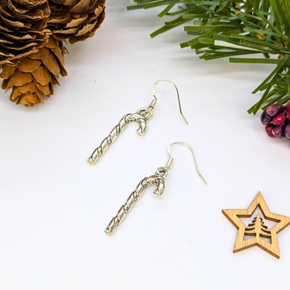 Two Classic Candy Cane Earrings by Silver Sunbird are displayed on a white surface with festive pine cones, pine needles, berries, and a star ornament—perfect Christmas jewelry for the holiday season.